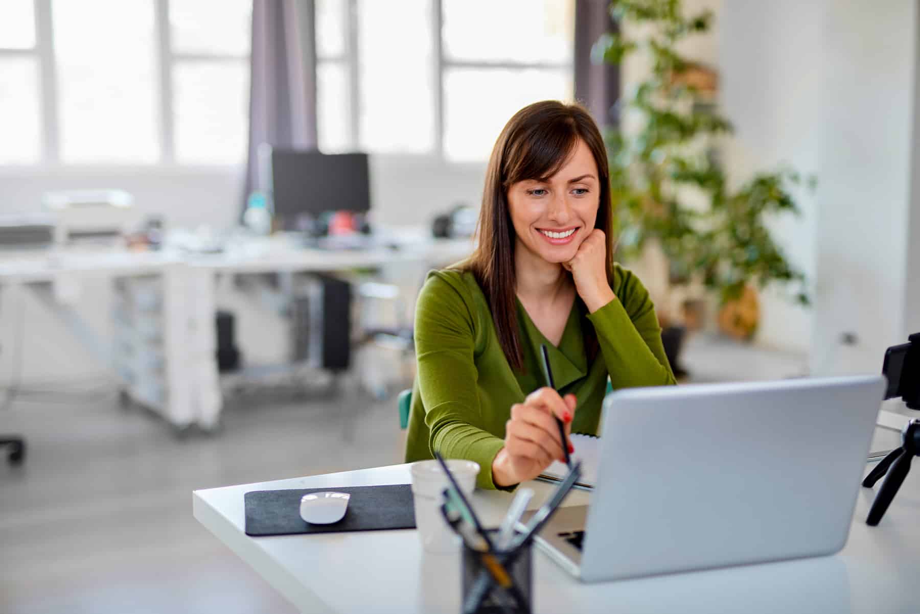 Woman holding pencil an looking at laptop while sitting in office.