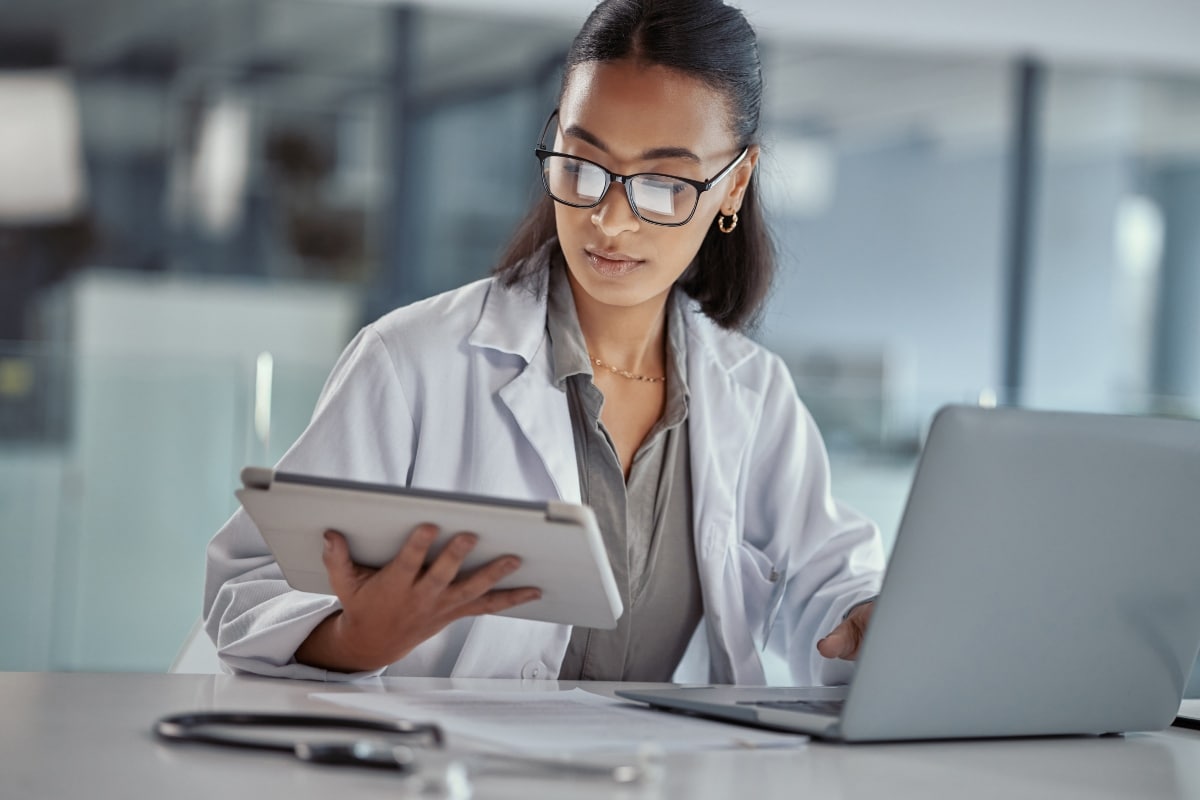 A lab technician compares duplicate patient records across her tablet and laptop.